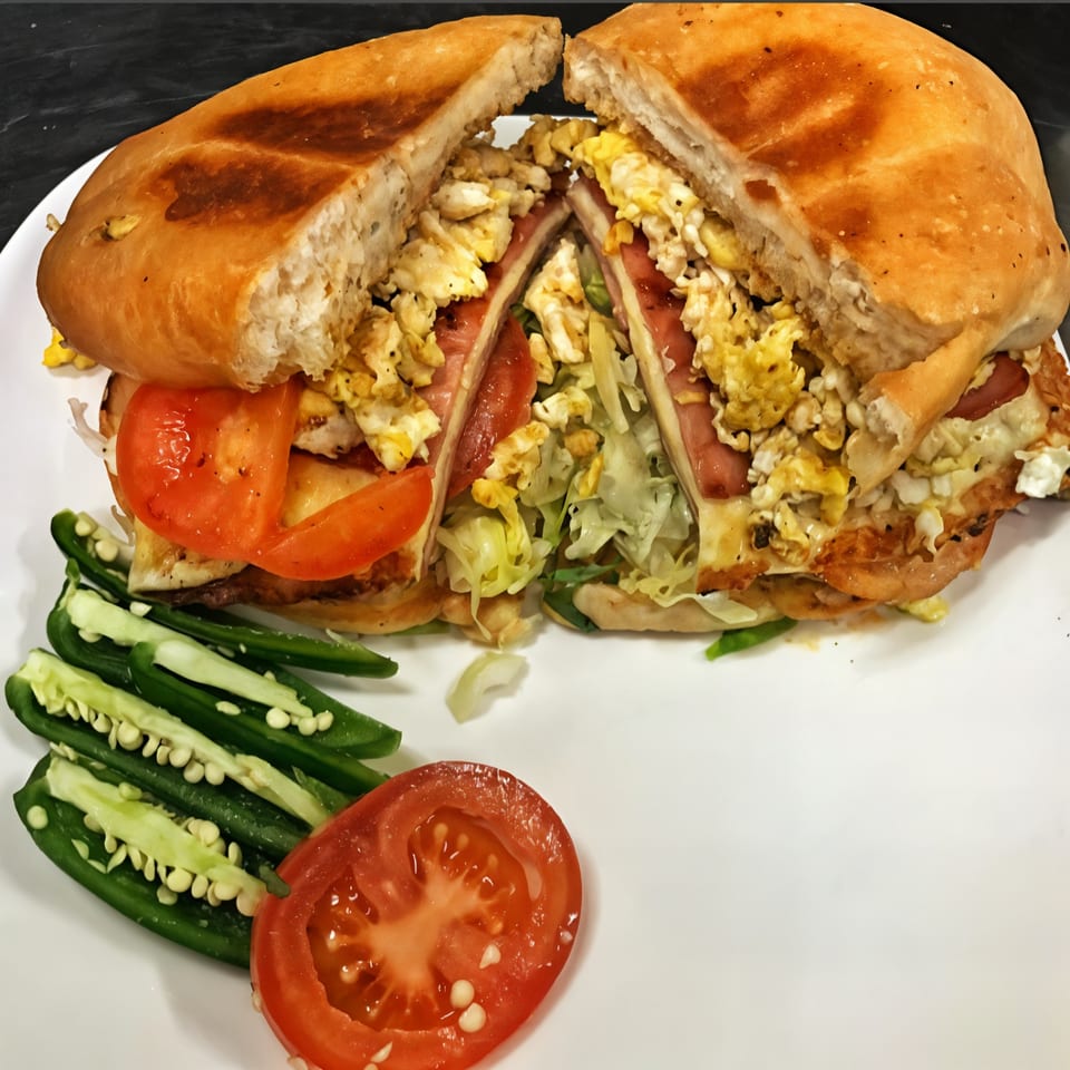 A Mexican torta sandwich, cut in half, filled with scrambled eggs, ham, lettuce, and tomato, served with sliced jalapeños.