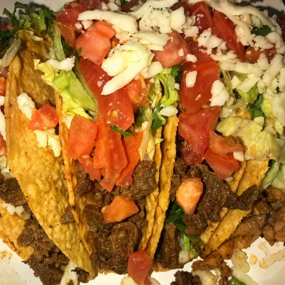Three crispy tacos from The Taco Place, filled with meat, lettuce, diced tomatoes, and shredded cheese.