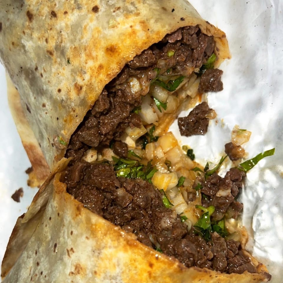 A large, cut open Mexican burrito from The Taco Place, filled with seasoned meat, onions, and cilantro.